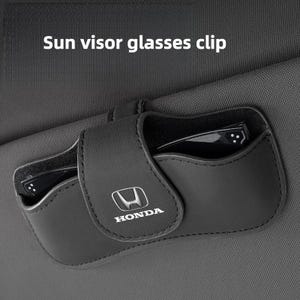 May include: A black leather-like sun visor glasses clip featuring the Honda logo on the front flap. The accessory is attached to a dark grey fabric car sun visor and contains a pair of black-framed glasses inside the storage compartment.