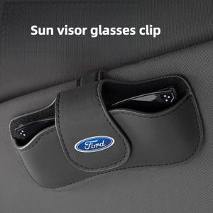 Könnte beinhalten: Ein schwarzer Sonnenblenden-Brillenhalter für das Auto mit dem Ford-Logo auf der Vorderklappe. Der Halter ist an einer dunkelgrauen Sonnenblende aus Stoff befestigt und enthält eine schwarze Brille. Der Text Sun visor glasses clip ist sichtbar.