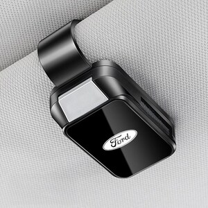 Könnte beinhalten: Ein schwarzer Sonnenbrillenhalter für die Sonnenblende im Auto mit glänzender Oberfläche und dem weißen Ford-Logo auf der Vorderseite. Das Zubehör verfügt über einen stabilen Befestigungsclip zur sicheren Anbringung an der Sonnenblende.