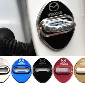 May include: A set of five metallic Mazda door latch covers in silver, blue, black, red, and gold. Each shield-shaped cover features the Mazda logo and text, with a central horizontal cutout designed to fit over a vehicle door striker mechanism.