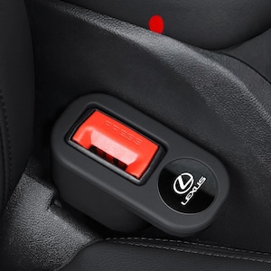 Puede incluir: Una funda negra para hebilla de cinturón de seguridad de coche con un botón de liberación rojo brillante con la palabra PRESS en relieve. La funda incluye un emblema circular negro con el logotipo y el texto de Lexus en color blanco.