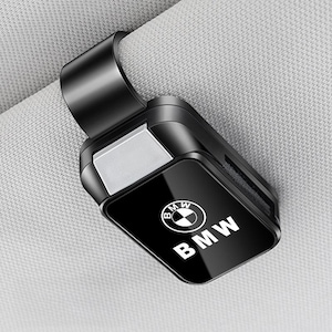 May include: A black plastic car visor sunglasses holder clip featuring a glossy front panel with the white BMW logo and text. The rectangular accessory has a spring-loaded mechanism designed to attach securely to a vehicle sun visor.