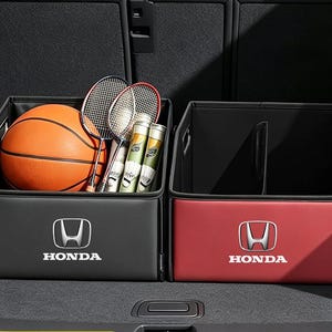 Puede incluir: Dos organizadores de almacenamiento rectangulares con el logotipo de Honda impreso en el frente. Uno es negro y contiene un balón de baloncesto naranja, dos raquetas de bádminton y dos tubos de volantes. El otro es rojo y está vacío.