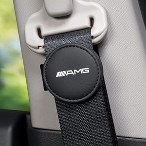 Könnte beinhalten: Ein schwarzer, runder Sicherheitsgurt-Clip mit weißem AMG-Logo, befestigt an einem schwarzen Standard-Sicherheitsgurtband. Das Zubehör befindet sich in der Nähe des hellgrauen Kunststoff-Höhenverstellmechanismus im Fahrzeuginnenraum.