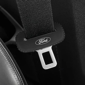 Op de afbeelding: Een zwarte veiligheidsgordel met een zwarte beschermhoes waarop het witte Ford-logo aan de voorzijde is gedrukt. De hoes is rechthoekig met afgeronde randen en bevindt zich boven de zilverkleurige metalen gesp van het veiligheidsgordelsysteem.
