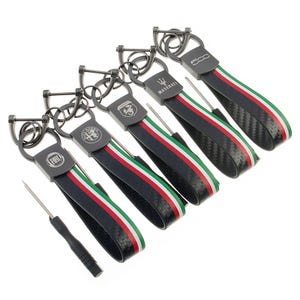 Puede incluir: Una colección de llaveros con correas negras y detalles metálicos. Cada llavero presenta un logotipo de marca de automóvil diferente, incluyendo Fiat, Alfa Romeo, Maserati y 500. Las correas tienen rayas rojas, blancas y verdes.