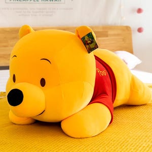 May include: A large, plush Winnie the Pooh stuffed animal in a lying position. The bear is yellow with a black nose and brown eyes, wearing a red shirt. A tag is attached to the ear.