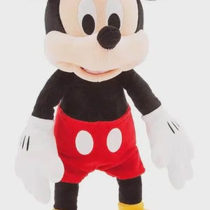 May include: A plush Mickey Mouse toy. The character has a black body, large black ears, and a white face. He wears red shorts with white buttons, white gloves, and yellow shoes. The toy is standing upright.