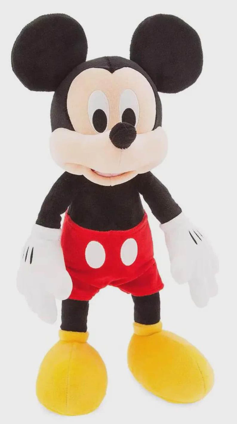 May include: A plush Mickey Mouse toy. The character has a black body, large black ears, and a white face. He wears red shorts with white buttons, white gloves, and yellow shoes. The toy is standing upright.