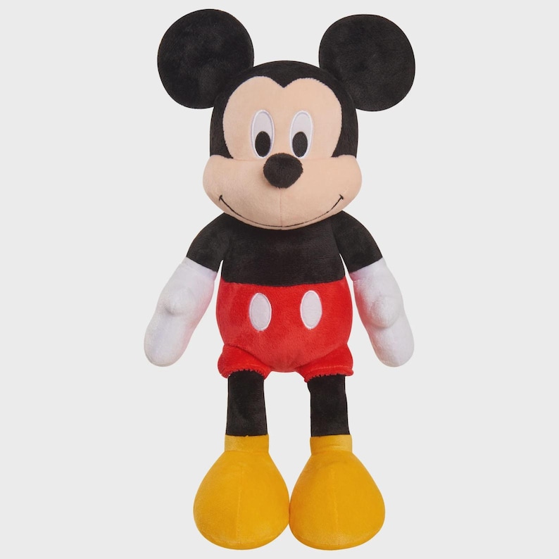 May include: A plush Mickey Mouse toy. The stuffed animal has a black body, large black ears, white gloves, red shorts with two white buttons, and yellow shoes. The toy is standing upright against a white background.