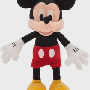 May include: A Mickey Mouse soft toy. The character has large black ears, a tan face, and a black body. He wears red shorts with white buttons, white gloves, and yellow shoes. The toy is designed to stand upright.