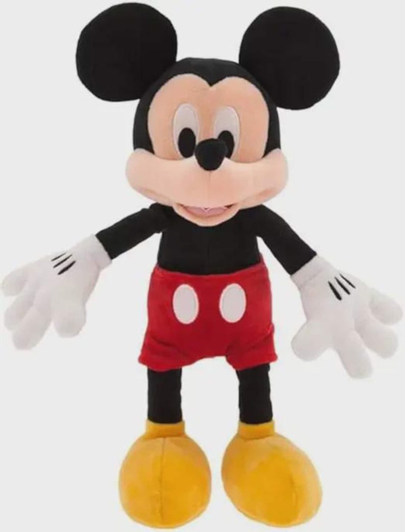 May include: A Mickey Mouse soft toy. The character has large black ears, a tan face, and a black body. He wears red shorts with white buttons, white gloves, and yellow shoes. The toy is designed to stand upright.