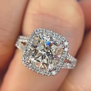 Puede incluir: Primer plano de un anillo de diamantes. El anillo presenta un diamante grande de talla cuadrada rodeado por un halo de diamantes más pequeños. La banda también está incrustada con diamantes, creando una apariencia brillante y lujosa.