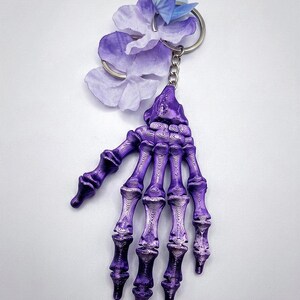 May include: A purple skeleton hand keychain featuring a metallic finish with visible 3D printed layer lines. The hand is attached to a silver metal split ring adorned with several light purple and blue artificial fabric flower petals.