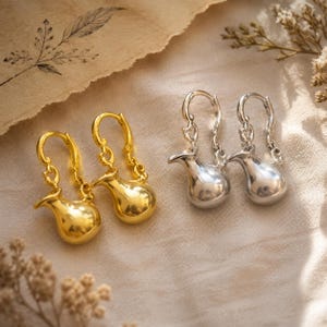 Peut inclure: Deux paires de boucles d'oreilles pendantes, une dorée et une argentée. Chaque boucle présente une petite breloque lisse et arrondie en forme de pichet, suspendue à un crochet incurvé par une courte chaîne délicate, créant un effet pendant.