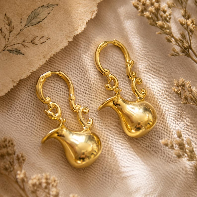 May include: A pair of gold-toned earrings featuring a polished, rounded base shaped like a small pitcher or vessel, connected to a decorative, curved wire hoop with intricate scrollwork details along the upper section of the earring structure.