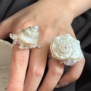 May include: Two rings featuring iridescent white seashell centrepieces. The left ring has a textured silver base with a spiral shell, while the right ring features a flat, circular shell with a silver wire spiral accent on the surface.