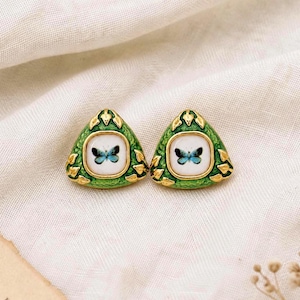 May include: A pair of triangular stud earrings featuring a gold-tone metal frame with green enamel detailing. Each earring has a central white square inlay decorated with a small blue butterfly graphic. The edges of the frame are textured with gold leaf motifs.