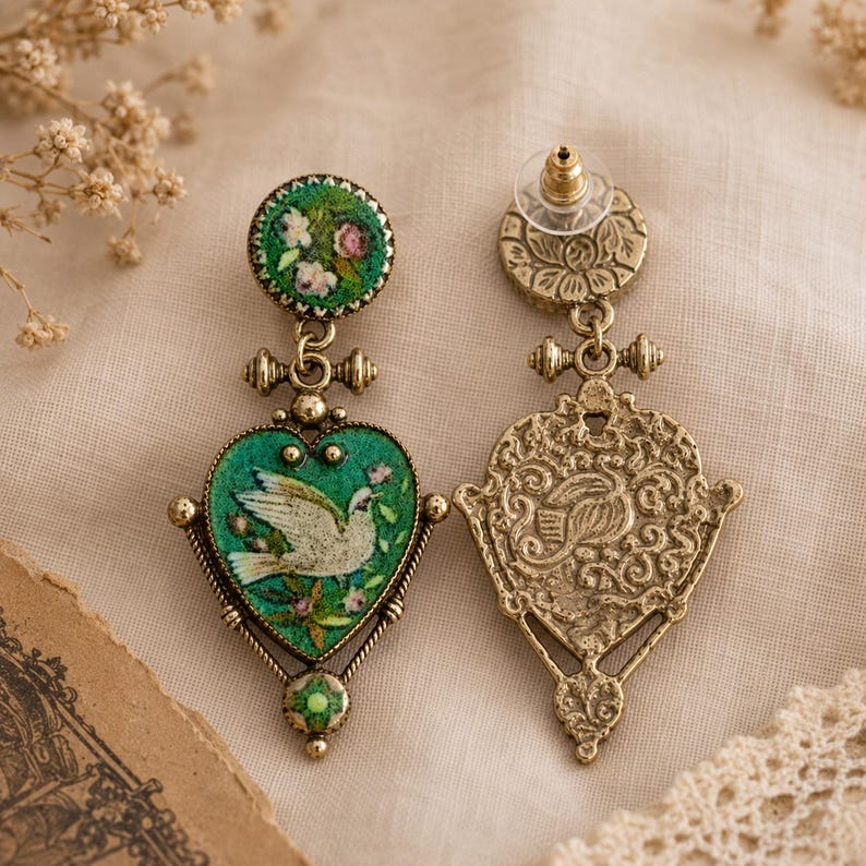 May include: A pair of drop earrings featuring an antique gold-tone metal finish. One earring displays a heart-shaped pendant with a green background, a white dove, and floral accents. The other earring shows the intricate, embossed metal back.