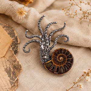 Op de afbeelding: Een zilverkleurige metalen broche met een inktviskop met meerdere krullende tentakels die omhoog steken, bevestigd aan een ronde, bruine, spiraalvormige ammoniet fossiele schelp met zichtbare gesegmenteerde kamers en een glad oppervlak.