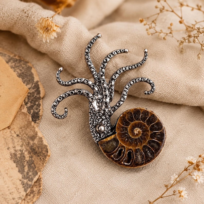 Op de afbeelding: Een zilverkleurige metalen broche met een inktviskop met meerdere krullende tentakels die omhoog steken, bevestigd aan een ronde, bruine, spiraalvormige ammoniet fossiele schelp met zichtbare gesegmenteerde kamers en een glad oppervlak.