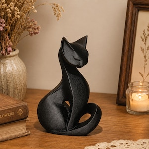 Puede incluir: Una figura de gato en color negro mate con una pose sentada y estilizada, la cabeza inclinada hacia abajo y los ojos cerrados. La escultura presenta líneas curvas y suaves con un espacio hueco entre el cuerpo y la cola, formando una pieza.