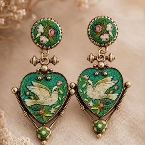 May include: A pair of drop earrings featuring a round top stud and a heart-shaped pendant, both in a textured green finish with floral accents. The heart pendants display a white dove illustration. The metal components are a muted gold tone.