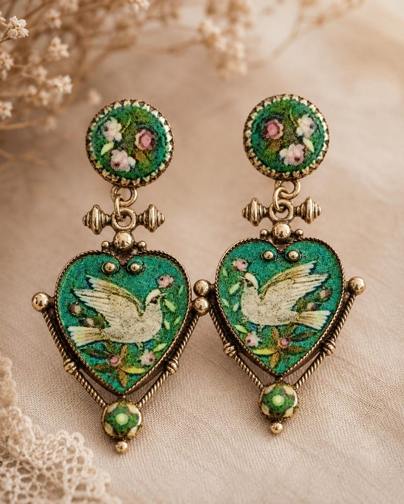 May include: A pair of drop earrings featuring a round top stud and a heart-shaped pendant, both in a textured green finish with floral accents. The heart pendants display a white dove illustration. The metal components are a muted gold tone.