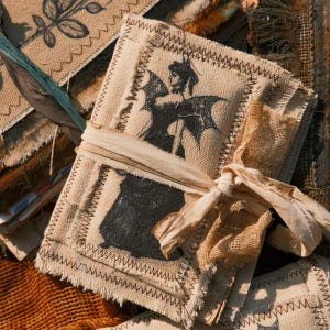 Op de afbeelding: Een rechthoekig dagboek ingebonden in beige jute stof met gerafelde randen en donkere zigzagstiksels. De kaft is voorzien van een zwarte zeefdruk van een gevleugeld figuur. Een bijpassend beige stoffen lint is rond het midden van het boek gebonden.