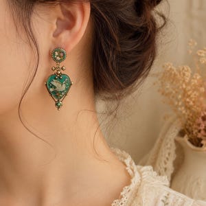 May include: A drop earring featuring a round stud with a floral pattern and a heart-shaped pendant. The pendant has a teal background with a white dove illustration, framed in antique gold-tone metal with small green accents at the base.