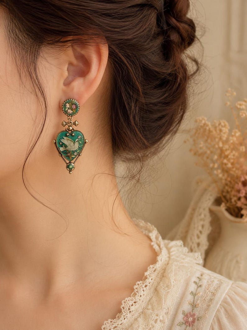 May include: A drop earring featuring a round stud with a floral pattern and a heart-shaped pendant. The pendant has a teal background with a white dove illustration, framed in antique gold-tone metal with small green accents at the base.