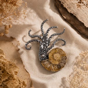 Op de afbeelding: Een broche met een zilverkleurige metalen inktviskop met meerdere gebogen tentakels die omhoog steken, bevestigd aan een ronde, goudbruine gefossiliseerde ammonietschelp met zichtbare spiraalkamers en een textuurrijk, kristallijn oppervlak.