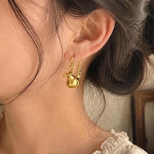May include: A gold-toned earring featuring a small, rounded, pitcher-shaped pendant suspended from a curved hook. The pendant has a smooth, reflective surface with a handle and a spout, hanging from a delicate, ornate metal loop attachment.