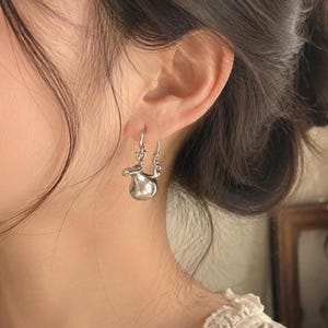 May include: A silver-toned drop earring featuring a small, smooth, bulbous pendant with an organic, asymmetrical shape. The pendant hangs from a simple metal hook that passes through the earlobe, showcasing a polished, reflective metallic surface.