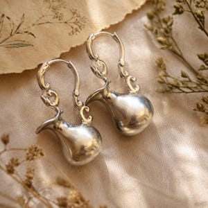 May include: A pair of silver-toned hoop earrings featuring a dangling, polished, bulbous pitcher-shaped charm. The upper portion of each earring consists of an ornate, curved metal structure that connects to a hinged hoop closure mechanism.