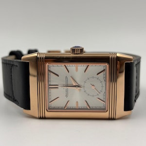 Op de afbeelding: Een rechthoekig roségouden polshorloge met een zilveren wijzerplaat en een zwarte leren band. De wijzerplaat heeft de merknaam en een kleine subwijzerplaat. Het horloge heeft een kroon met een blauwe steen.