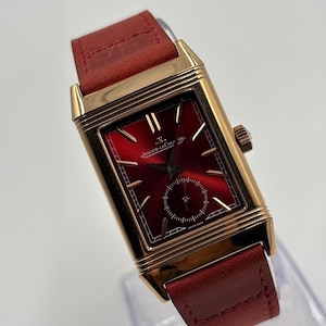 May include: A rectangular gold-toned wristwatch with a burgundy dial and a small seconds subdial. The watch features a burgundy leather strap. The watch face has the text "JAEGER-LECOULTRE" and "SWISS MADE".