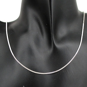 May include: A delicate silver necklace with a box chain design. The necklace is displayed on a black mannequin, showcasing its simple elegance. The chain is thin and appears to be made of a shiny metal.