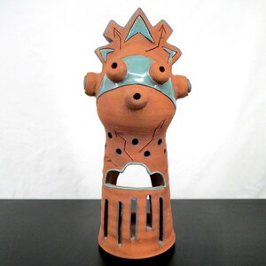May include: A handcrafted ceramic sculpture in shades of orange and teal. The whimsical piece features a stylized face with large, round eyes and a decorative crown. The body has cut-out details and a cylindrical base.