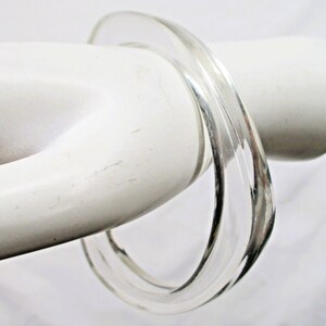 May include: A clear, transparent bangle bracelet. The bracelet is made of a solid, smooth material and has a rounded shape. The light reflects off the surface, creating highlights and shadows. The bangle is a simple, elegant accessory.