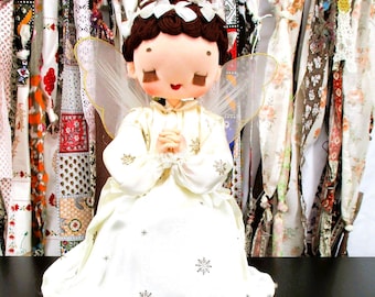 May include: A fabric angel figurine with brown hair, closed eyes, and a red mouth. The angel wears a cream-colored gown featuring a gold snowflake pattern, white ribbon bows in the hair, a pearl-beaded halo, and sheer white wings on the back.