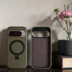 May include: Two olive green phone cases. One case has a circular magnetic attachment and a camera cutout. The other case is open, revealing a brown interior. The cases are displayed on a dark surface with a vase of flowers in the background.