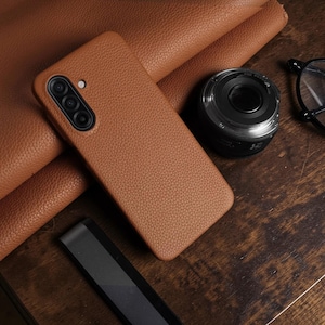 May include: A brown leather phone case with a textured surface. The case has a camera cutout with three lenses. A black camera lens and eyeglasses are also on the wooden surface.