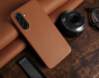Puede incluir: Una funda de teléfono de cuero marrón con una superficie texturizada. La funda tiene un recorte para la cámara con tres lentes. Una lente de cámara negra y gafas también están sobre la superficie de madera.