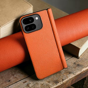 Puede incluir: Funda de teléfono de cuero naranja con un acabado texturizado. La funda tiene un módulo de cámara negro y un diseño plegable. La funda del teléfono está sobre una superficie de madera con un artículo de cuero enrollado y un libro en el fondo.