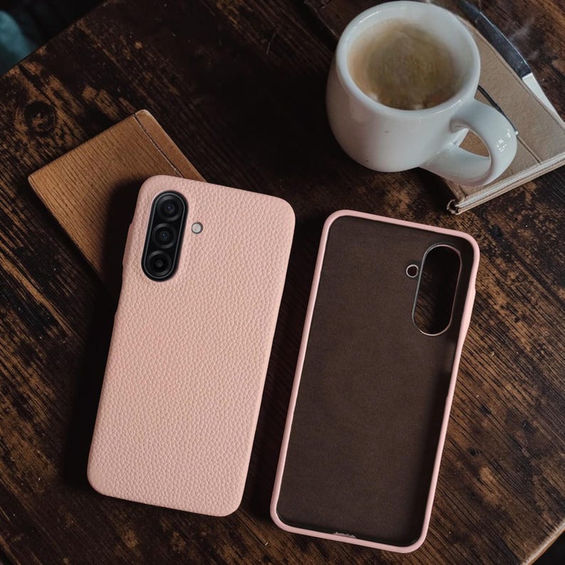 Op de afbeelding: Twee roze telefoonhoesjes, &eacute;&eacute;n gesloten en &eacute;&eacute;n open, liggen op een donker houten oppervlak. De gesloten hoes heeft een getextureerde afwerking en een camera-array. De open hoes onthult een bruin interieur. Een witte koffiemok en een klein notitieboekje staan op de achtergrond.