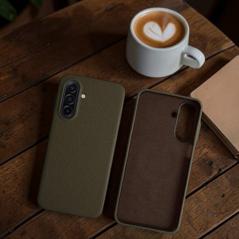 Op de afbeelding: Twee olijfgroene telefoonhoesjes, waarvan &eacute;&eacute;n open, met een bruin interieur, op een houten oppervlak. Een kopje koffie met een hartvormig patroon in het schuim staat naast de hoesjes. Een notitieboek is ook zichtbaar.
