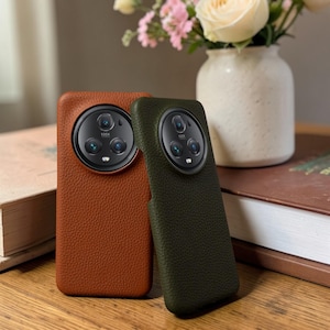May include: Two leather phone cases, one in a warm brown and the other in a deep olive green, are displayed. The cases have a textured surface and a circular camera cutout with the text "100X". A vase of flowers sits in the background.