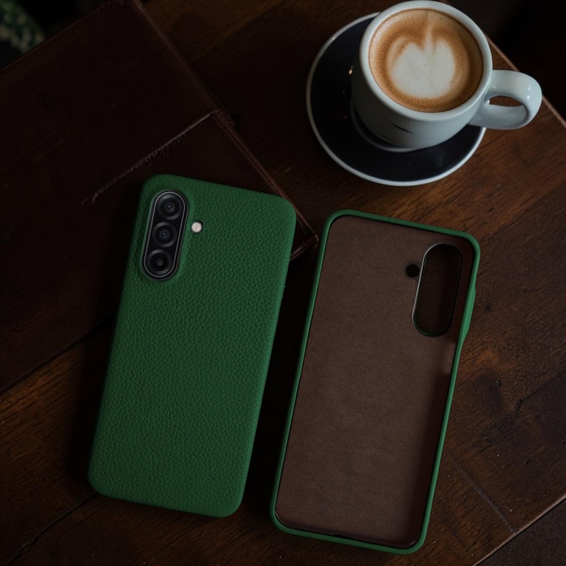 Op de afbeelding: Twee groene telefoonhoesjes, &eacute;&eacute;n open en &eacute;&eacute;n gesloten, liggen op een houten oppervlak. De gesloten hoes heeft een getextureerde afwerking en een camera-opstelling. Een kopje koffie met latte art staat op de achtergrond.
