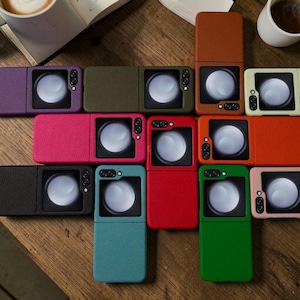 May include: A collection of colorful phone cases in various shades, including purple, pink, green, and red. Each case features a camera cutout and a textured surface, arranged on a wooden surface. The cases are designed to fit a specific phone model.